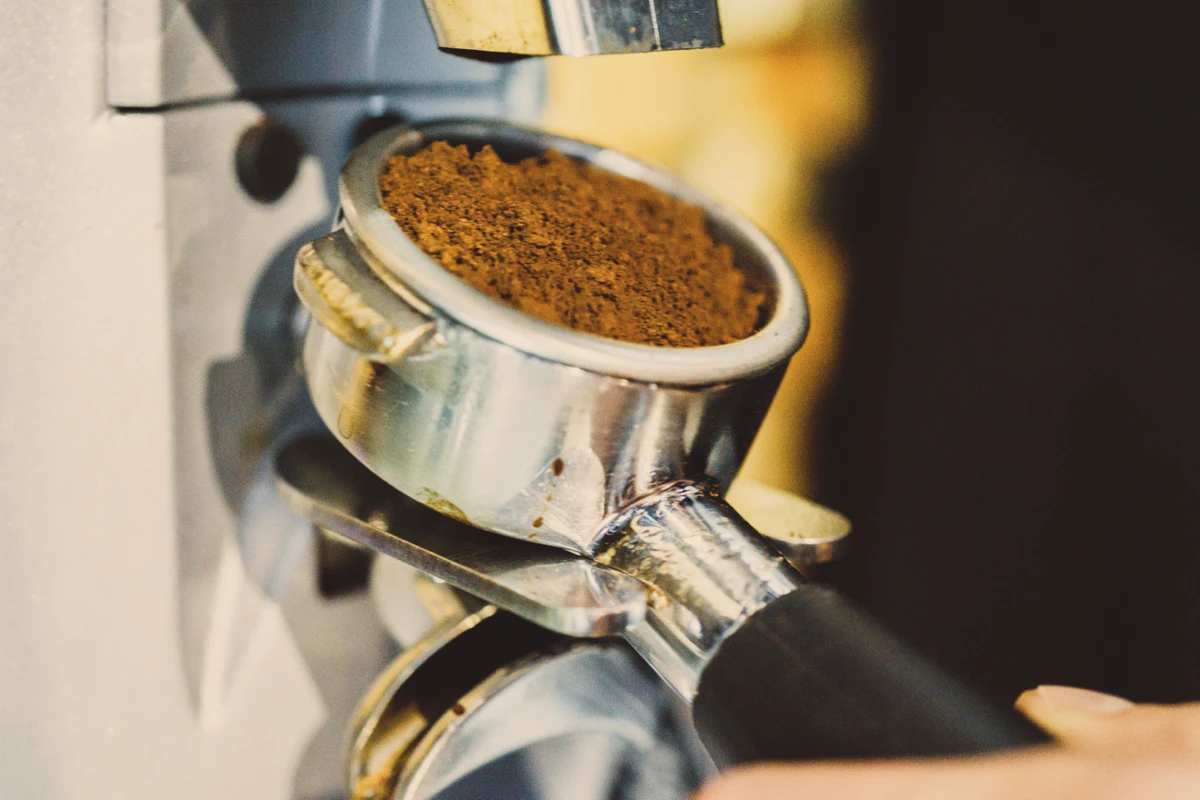 Close-up of a portafilter filled with ground coffee being held up to a coffee grinder.