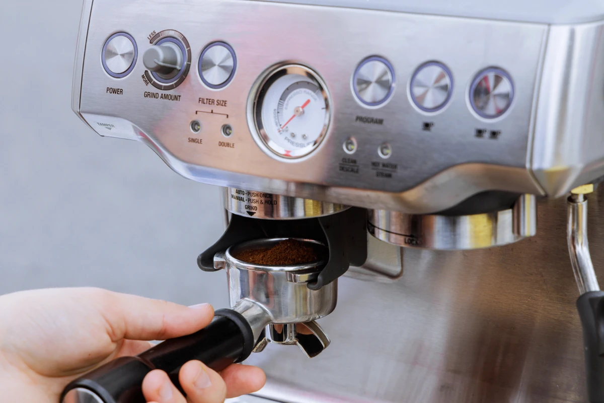 Close-up of a hand holding a portafilter with ground coffee under the brew head of an espresso machine with visible buttons and pressure gauge.