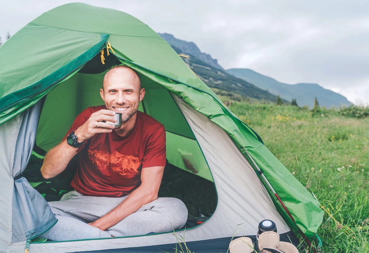A man sits cross-legged in a green tent, holding a metal cup and smiling, with mountains and grass visible in the background.