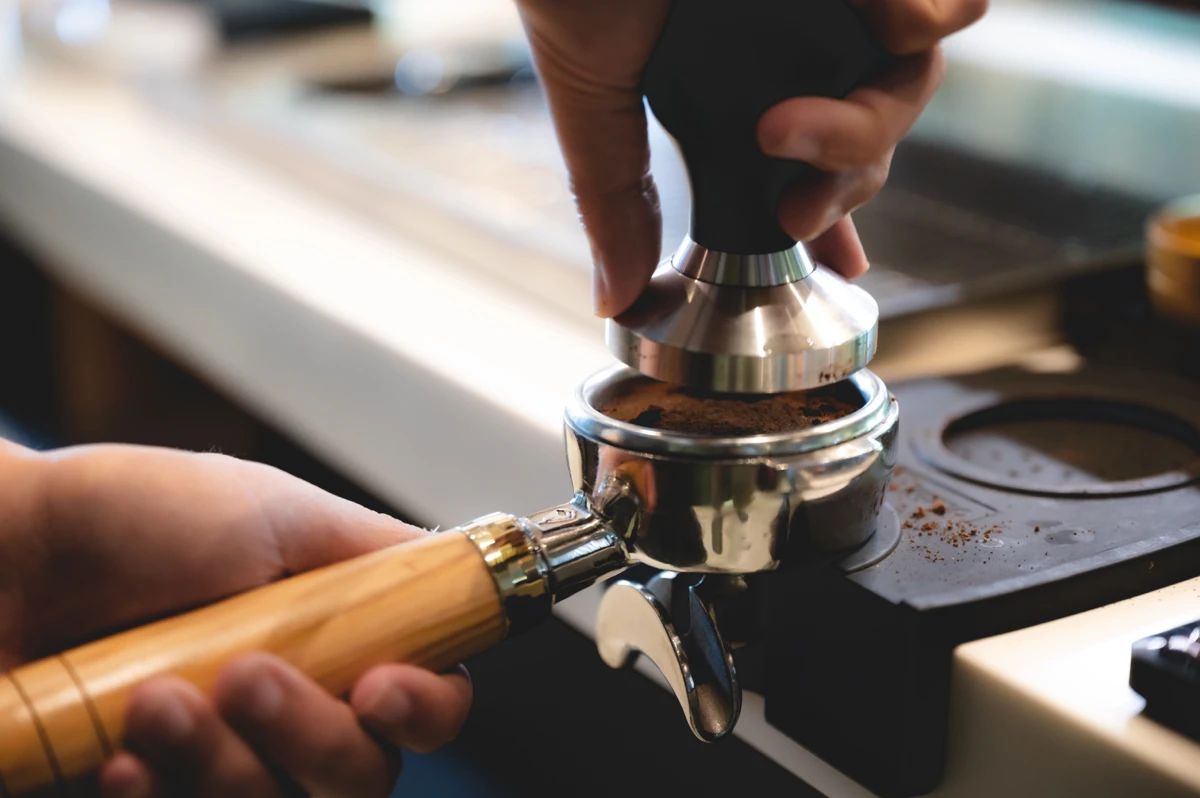 A person uses a tamper to press ground coffee into a portafilter, preparing it for espresso brewing on a countertop. A person uses a tamper to press ground coffee into a portafilter, preparing it for espresso brewing on a countertop.