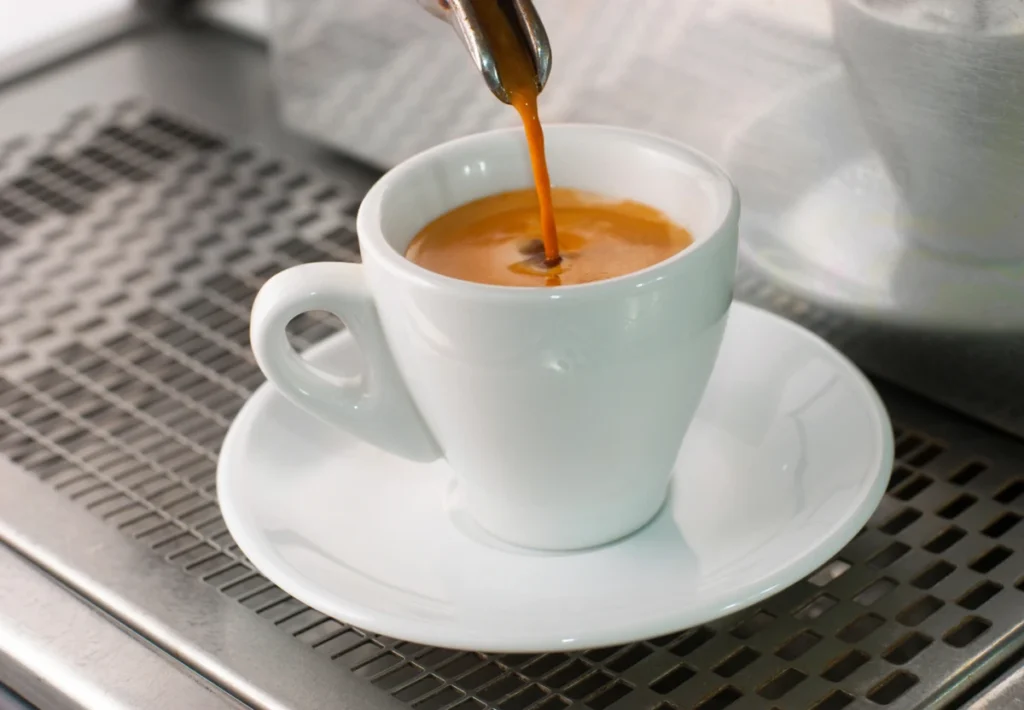 A shot of espresso is being poured into a white cup on a saucer, placed on an espresso machine drip tray.