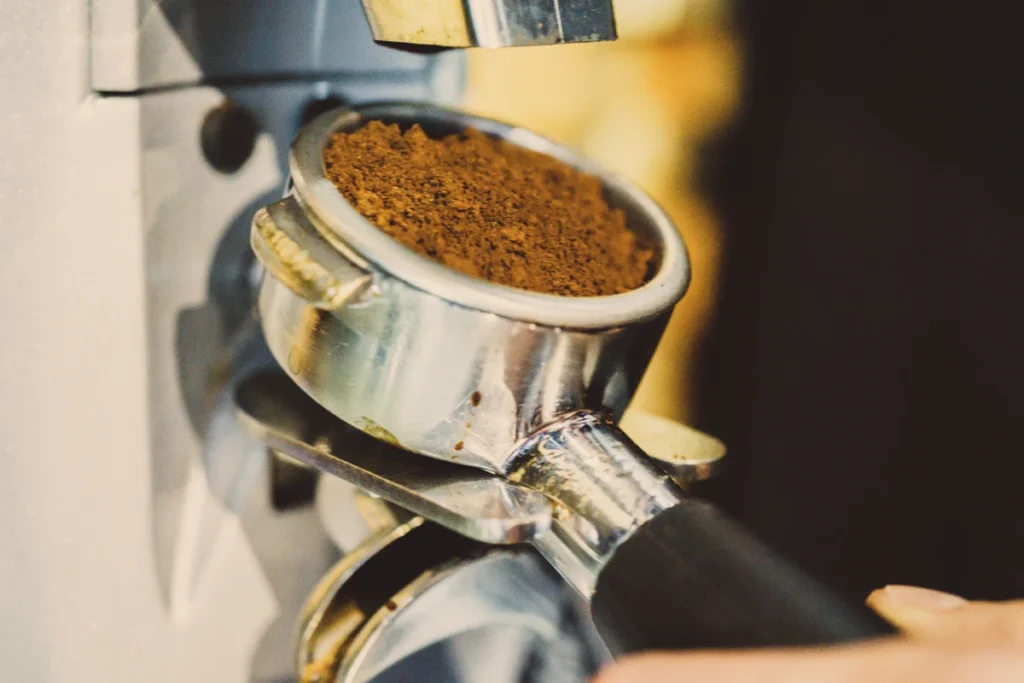 Close-up of a portafilter filled with ground coffee being held up to a coffee grinder.