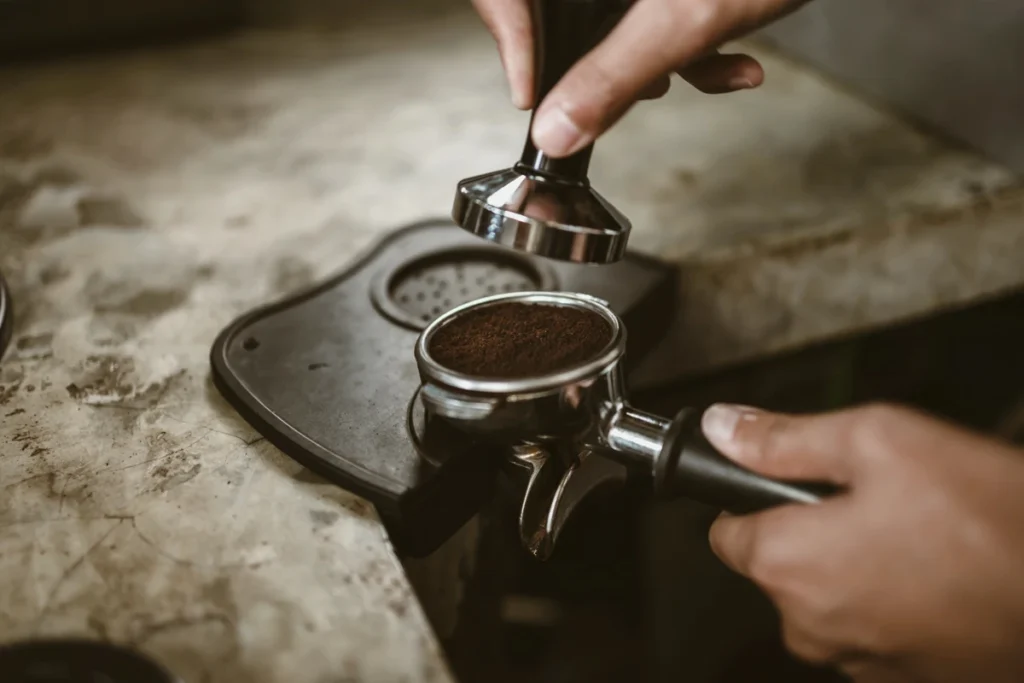 A person compresses ground coffee in a portafilter with a tamper on a countertop.