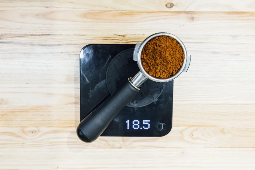 A portafilter filled with ground coffee sits on a digital scale displaying a weight of 18.5 grams, on a light wooden surface.