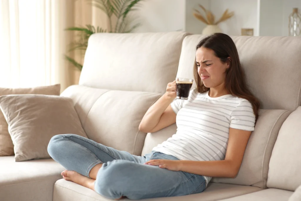 A woman sits on a beige sofa holding a mug, making a displeased face as she looks at her drink.