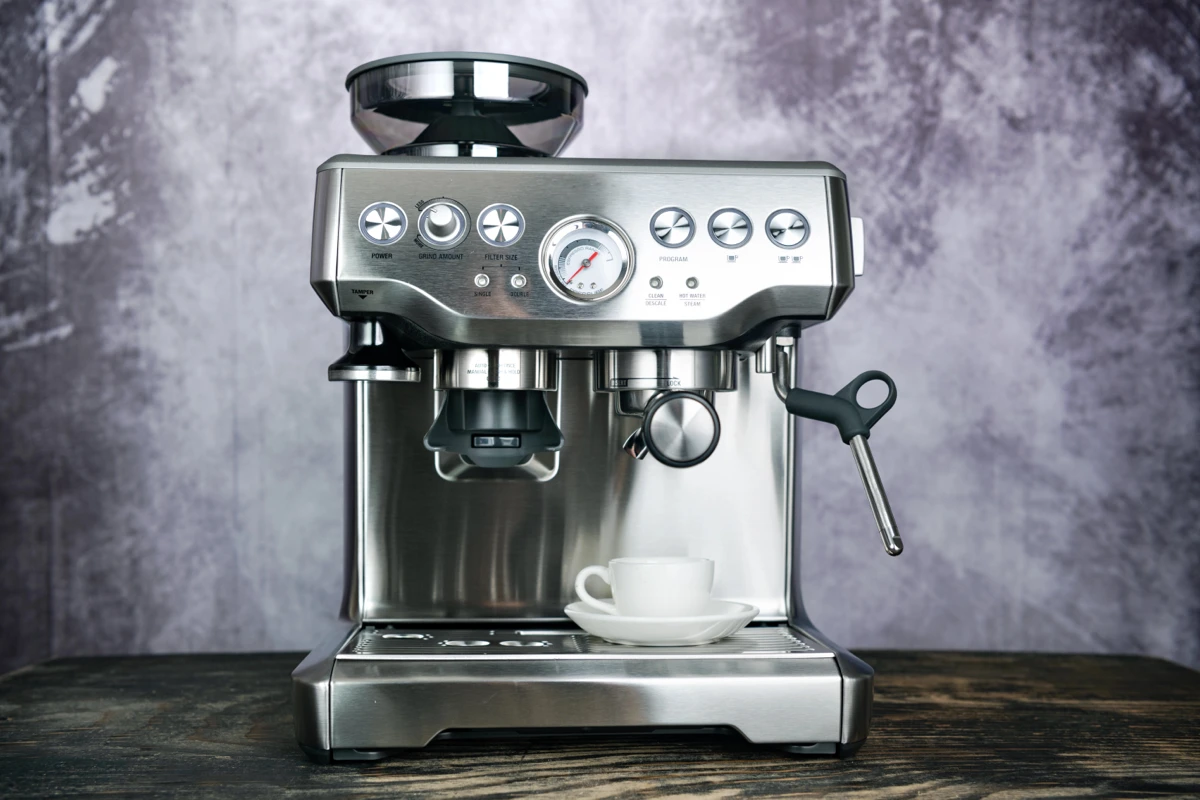 A stainless steel espresso machine with buttons and a gauge sits on a wooden surface, with a white cup and saucer placed on its drip tray.