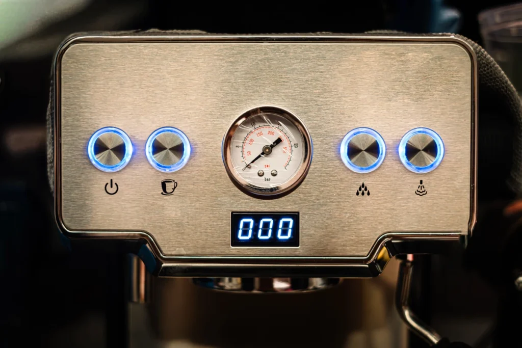 Close-up of a coffee machine control panel with four illuminated buttons, a pressure gauge in the center, and a digital display showing "000".