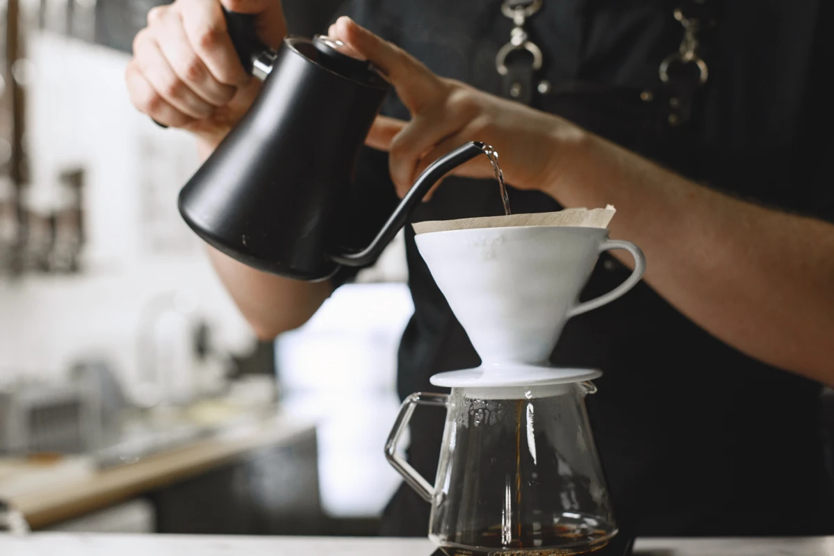 Person pouring hot water from a black kettle into a filter over a glass coffee carafe, making pour-over coffee.